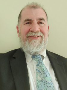 Image of Dr. Gary Schmidt smiling at the camera. He has a gray beard and wears a dark gray suit with a light blue paisley tie.