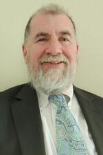 Image of Dr. Gary Schmidt smiling at the camera. He has a gray beard and wears a dark gray suit with a light blue paisley tie.